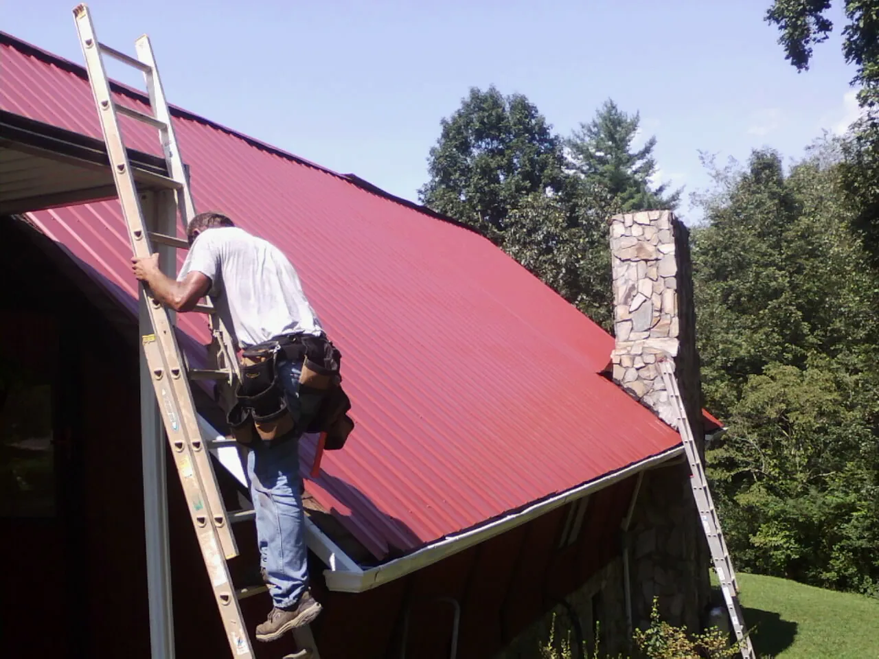 Roofer on a ladder beside a red metal roof and stone chimney — Soffit & Fascia Repair by Roofing Brothers in Huntingburg
